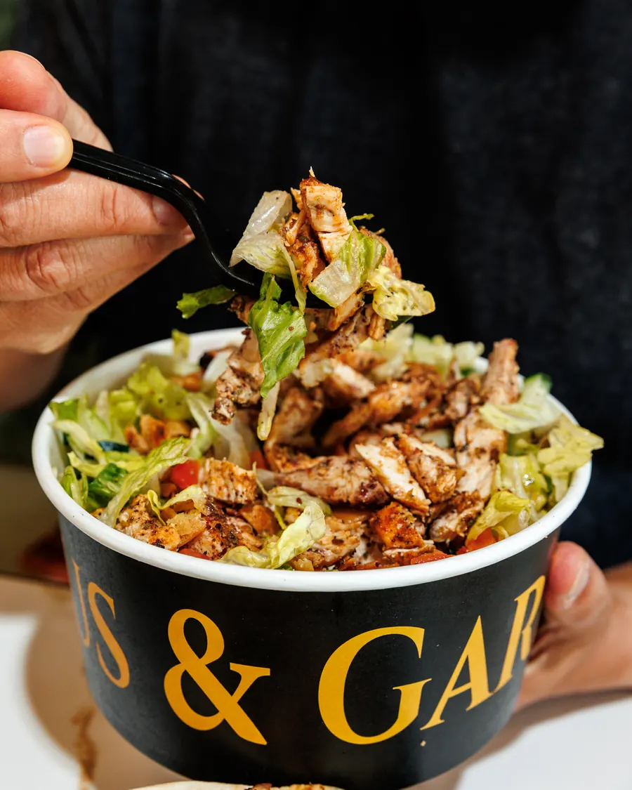 A fork lifting grilled chicken, lettuce and rice out of a Hummus & Garlic bowl
