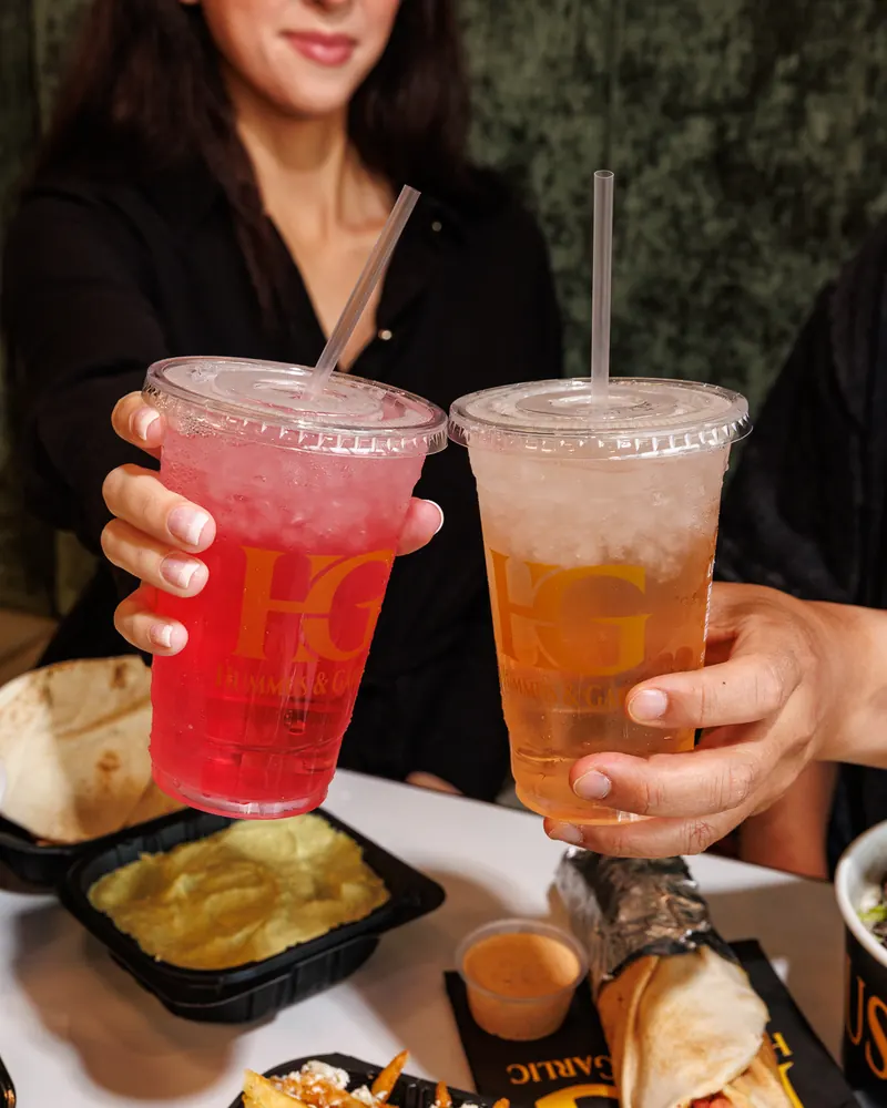 Two guests toasting with a strawberry lemonade and an iced tea over a table of shawarma and fries