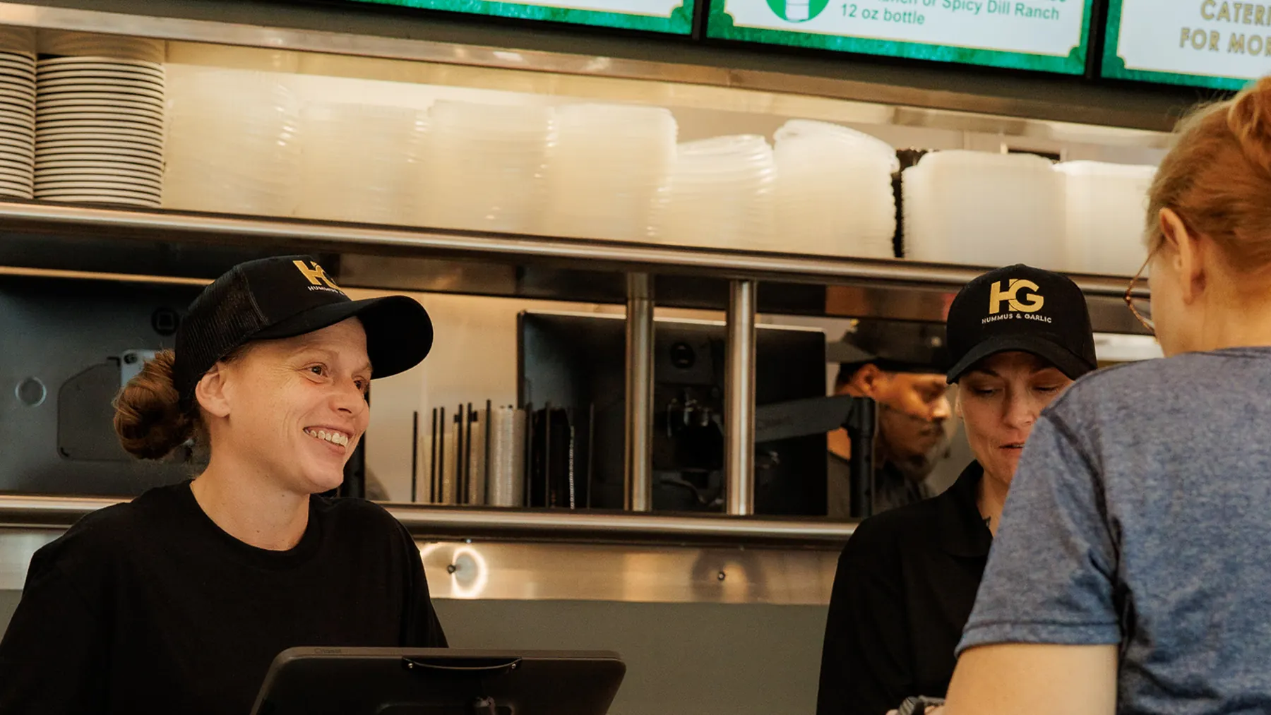Two smiling Hummus & Garlic team members in black caps serving a guest at the counter